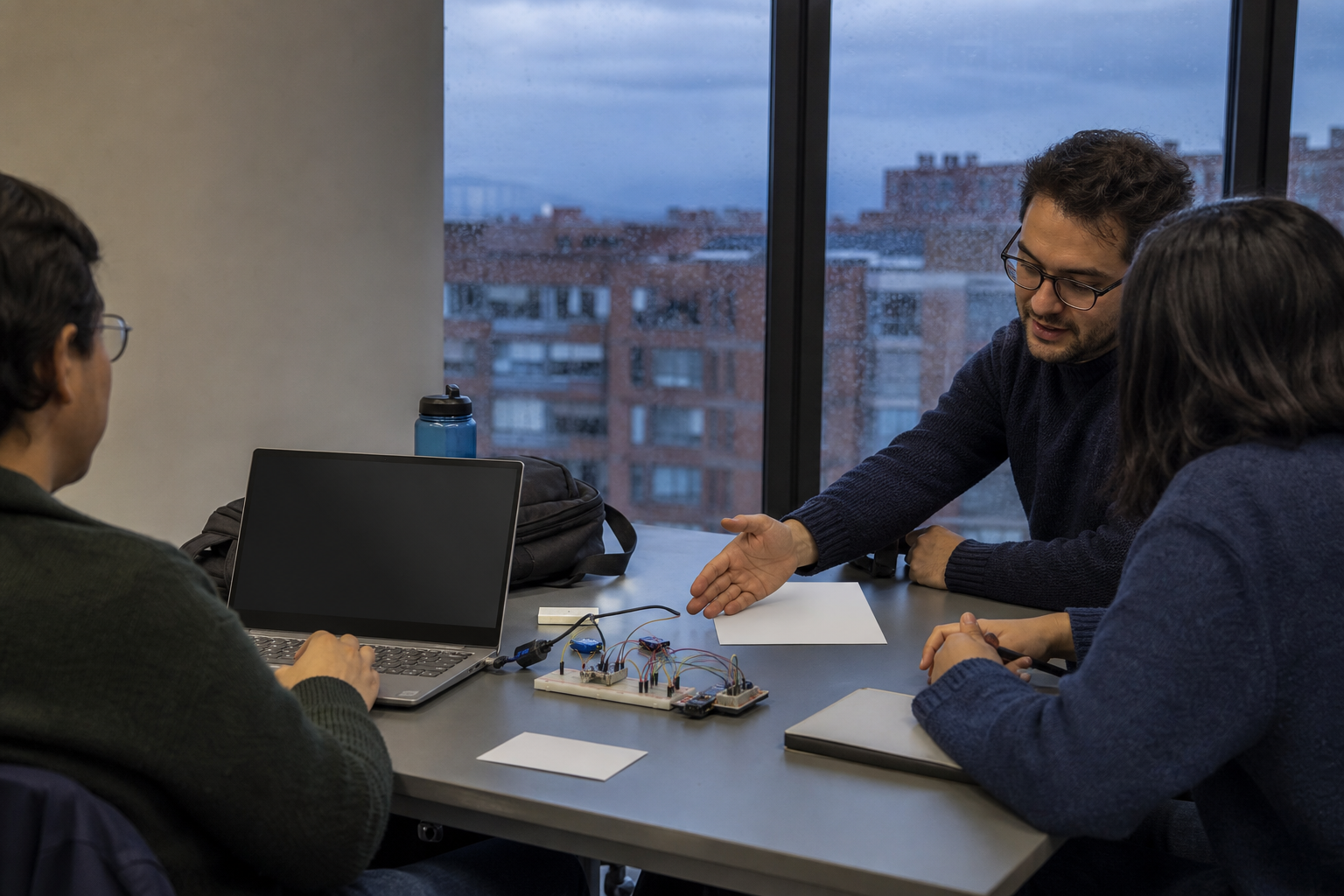 Escena editorial conceptual de estudiantes adultos aprendiendo inteligencia artificial en un aula de Bogotá, con computadores y un kit de sensores junto a una ventana lluviosa.